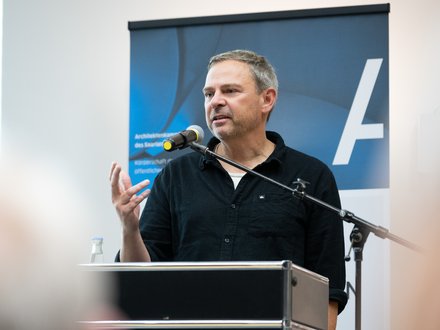 Mann mittleren Alters steht hinter einem Rednerpult und spricht ins Mikrofon; er trägt ein dunkles Hemd, gestikuliert mit der rechten Hand und blickt leicht nach rechts; hinter ihm ein blaues Roll‑up mit weißem Schriftzeichen als Hintergrund.