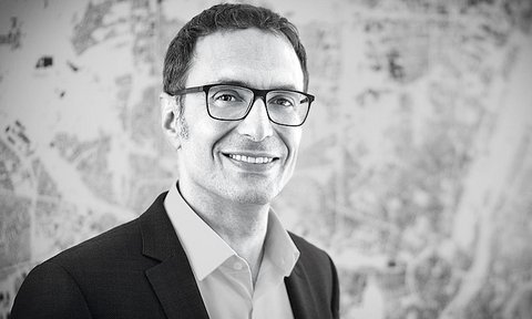 "Schwarzweiß-Porträtfoto eines Mannes mittleren Alters mit dunklem Haar und Brille. Er lächelt freundlich zur Kamera. Er trägt einen dunklen Blazer über einem hellen Hemd. Im unscharfen Hintergrund sind Skizzen oder Pläne erkennbar. Professionelle Headshot-Fotografie."