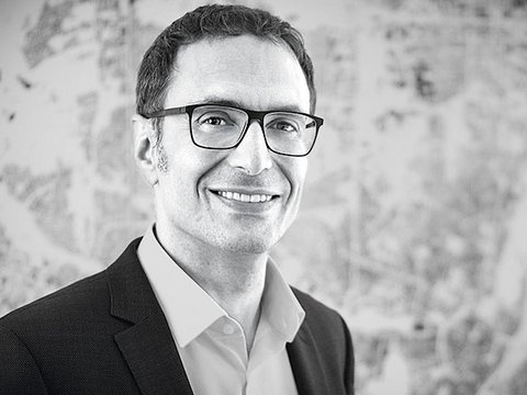 "Schwarzweiß-Porträtfoto eines Mannes mittleren Alters mit dunklem Haar und Brille. Er lächelt freundlich zur Kamera. Er trägt einen dunklen Blazer über einem hellen Hemd. Im unscharfen Hintergrund sind Skizzen oder Pläne erkennbar. Professionelle Headshot-Fotografie."