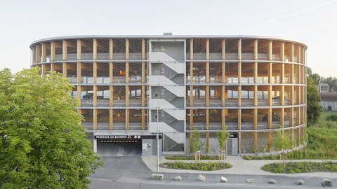 Tagesansicht eines runden Holzparkhauses mit offener Holzfassade, zentralem Treppenhaus und Zufahrt im Erdgeschoss, umgeben von Grünflächen.