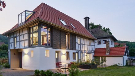 "Modernes Wohnhaus in der Abenddämmerung mit traditionellem Fachwerkstil. Das Gebäude hat ein rotes Satteldach mit Dachfenstern, weiße Fassade mit schwarzen Fachwerkbalken an der oberen Etage und große verglaste Fenster. Ein moderner weißer Anbau mit Glasdach befindet sich auf der linken Seite. Im Vordergrund grüne Rasenfläche mit Trittsteinen, rote Gartenstühle auf der Terrasse."