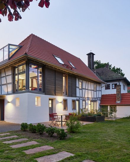 "Modernes Wohnhaus in der Abenddämmerung mit traditionellem Fachwerkstil. Das Gebäude hat ein rotes Satteldach mit Dachfenstern, weiße Fassade mit schwarzen Fachwerkbalken an der oberen Etage und große verglaste Fenster. Ein moderner weißer Anbau mit Glasdach befindet sich auf der linken Seite. Im Vordergrund grüne Rasenfläche mit Trittsteinen, rote Gartenstühle auf der Terrasse."