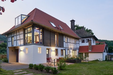 "Modernes Wohnhaus in der Abenddämmerung mit traditionellem Fachwerkstil. Das Gebäude hat ein rotes Satteldach mit Dachfenstern, weiße Fassade mit schwarzen Fachwerkbalken an der oberen Etage und große verglaste Fenster. Ein moderner weißer Anbau mit Glasdach befindet sich auf der linken Seite. Im Vordergrund grüne Rasenfläche mit Trittsteinen, rote Gartenstühle auf der Terrasse."