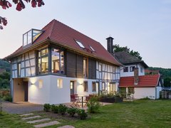 "Modernes Wohnhaus in der Abenddämmerung mit traditionellem Fachwerkstil. Das Gebäude hat ein rotes Satteldach mit Dachfenstern, weiße Fassade mit schwarzen Fachwerkbalken an der oberen Etage und große verglaste Fenster. Ein moderner weißer Anbau mit Glasdach befindet sich auf der linken Seite. Im Vordergrund grüne Rasenfläche mit Trittsteinen, rote Gartenstühle auf der Terrasse."