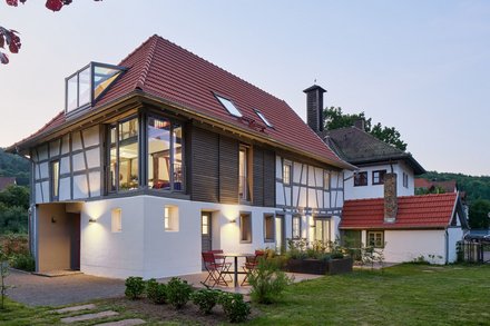 "Modernes Wohnhaus in der Abenddämmerung mit traditionellem Fachwerkstil. Das Gebäude hat ein rotes Satteldach mit Dachfenstern, weiße Fassade mit schwarzen Fachwerkbalken an der oberen Etage und große verglaste Fenster. Ein moderner weißer Anbau mit Glasdach befindet sich auf der linken Seite. Im Vordergrund grüne Rasenfläche mit Trittsteinen, rote Gartenstühle auf der Terrasse."