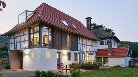 "Modernes Wohnhaus in der Abenddämmerung mit traditionellem Fachwerkstil. Das Gebäude hat ein rotes Satteldach mit Dachfenstern, weiße Fassade mit schwarzen Fachwerkbalken an der oberen Etage und große verglaste Fenster. Ein moderner weißer Anbau mit Glasdach befindet sich auf der linken Seite. Im Vordergrund grüne Rasenfläche mit Trittsteinen, rote Gartenstühle auf der Terrasse."