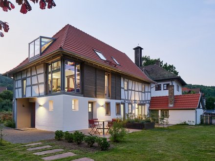 "Modernes Wohnhaus in der Abenddämmerung mit traditionellem Fachwerkstil. Das Gebäude hat ein rotes Satteldach mit Dachfenstern, weiße Fassade mit schwarzen Fachwerkbalken an der oberen Etage und große verglaste Fenster. Ein moderner weißer Anbau mit Glasdach befindet sich auf der linken Seite. Im Vordergrund grüne Rasenfläche mit Trittsteinen, rote Gartenstühle auf der Terrasse."