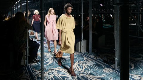 "Laufsteg einer Modenschau in einer industriellen Halle mit schwarzen Metallgitterstrukturen und Deckenbeleuchtung. Der Laufsteg ist mit gemustertem Stoff ausgelegt. Drei Models laufen einzeln über den Laufsteg und präsentieren feminine Kleidungsstücke in zartem Pink, Navy und Beige mit Accessoires. Der Publikumsbereich ist dunkel und umgibt den beleuchteten Laufsteg. Das Metallgerüst rahmt die Modenpräsentation ein."