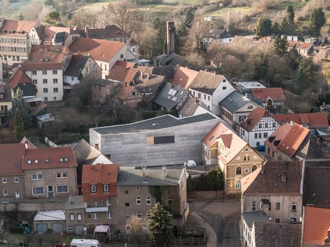"Vogelschau eines ländlichen Dorfes in einem Tal. Im Vordergrund ein modernes Gebäude mit grauer Betonoptik und großem dunklem Dach, umgeben von traditionellen Wohnhäusern mit roten und braunen Dächern. Links mehrgeschossige Mehrfamilienhäuser, rechts ein gelbes Fachwerkgebäude. Im Hintergrund weitläufige grüne Wiesen mit vereinzelten Bäumen und einem Schornstein. Der Himmel ist blau und klar."