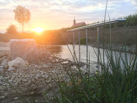 "Sonnenaufgang über einem Fluss mit Kiesstrand. Im Vordergrund grüne Schilfpflanzen und helle Kieselsteine. Eine moderne Steganlage mit Metallkonstruktion führt ins Wasser. Im Hintergrund eine Kirche mit rötlichem Dach auf einem Hügel, umgeben von Wald und Bäumen. Der Himmel zeigt Pastell-Orange und Rosa-Töne während der Sonnenaufgang die Landschaft in warmes Licht taucht."