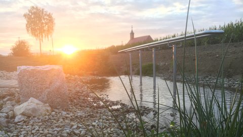 "Sonnenaufgang über einem Fluss mit Kiesstrand. Im Vordergrund grüne Schilfpflanzen und helle Kieselsteine. Eine moderne Steganlage mit Metallkonstruktion führt ins Wasser. Im Hintergrund eine Kirche mit rötlichem Dach auf einem Hügel, umgeben von Wald und Bäumen. Der Himmel zeigt Pastell-Orange und Rosa-Töne während der Sonnenaufgang die Landschaft in warmes Licht taucht."