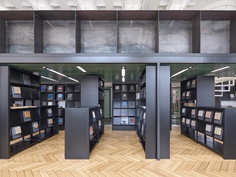 Eine Ansicht eines bibliotheksähnlichen Raums mit schwarzen Regalen, die verschiedene Materialproben halten, wobei natürliches Licht durch große Fenster strömt.
