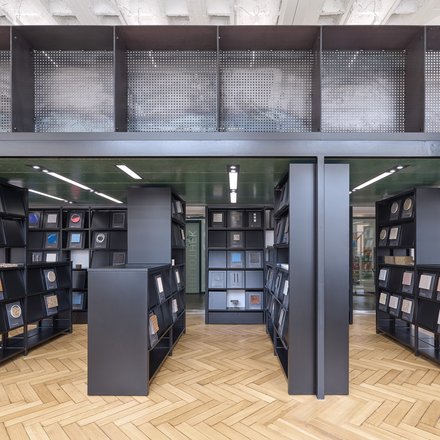 Eine Ansicht eines bibliotheksähnlichen Raums mit schwarzen Regalen, die verschiedene Materialproben halten, wobei natürliches Licht durch große Fenster strömt.