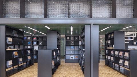 Eine Ansicht eines bibliotheksähnlichen Raums mit schwarzen Regalen, die verschiedene Materialproben halten, wobei natürliches Licht durch große Fenster strömt.