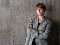 Porträt: Person mit kurzem braunen Haar im grauen Blazer steht mit verschränkten Armen vor einer rauen Betonwand und blickt ruhig in die Kamera.