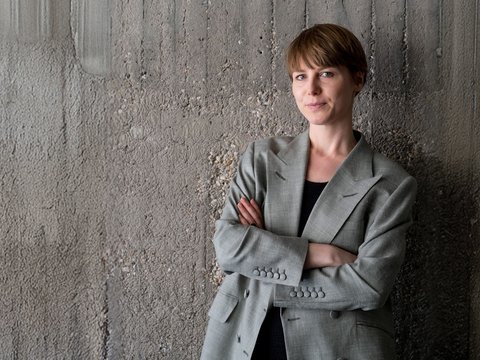 Porträt: Person mit kurzem braunen Haar im grauen Blazer steht mit verschränkten Armen vor einer rauen Betonwand und blickt ruhig in die Kamera.