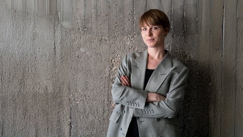 Porträt: Person mit kurzem braunen Haar im grauen Blazer steht mit verschränkten Armen vor einer rauen Betonwand und blickt ruhig in die Kamera.