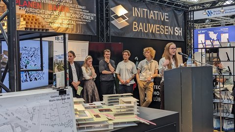 Eine Person spricht an einem Rednerpult vor einem Messestand, im Vordergrund sind Architekturmodelle und Pläne ausgestellt, im Hintergrund stehen mehrere junge Erwachsene und hören zu, große Banner mit Begriffen rund um Planen, Bauen und Nachhaltigkeit rahmen die Szene, wodurch das Bild eine Präsentation oder Projektvorstellung zeigt und für Wissensvermittlung, Nachwuchsförderung und den öffentlichen Austausch über zeitgenössische Baukultur steht.