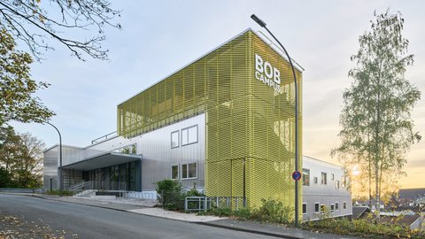 Modernes Gebäude mit markanter gelber, gitterartiger Fassade und der Aufschrift ‚BOB CAMPUS‘; silberne Gebäudeteile, Straßenlaterne und eine Birke rechts, leicht ansteigende Straße mit Laub, Sonne am Horizont