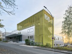 Modernes Gebäude mit markanter gelber, gitterartiger Fassade und der Aufschrift ‚BOB CAMPUS‘; silberne Gebäudeteile, Straßenlaterne und eine Birke rechts, leicht ansteigende Straße mit Laub, Sonne am Horizont