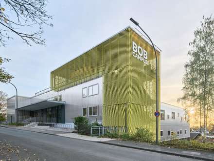 Modernes Gebäude mit markanter gelber, gitterartiger Fassade und der Aufschrift ‚BOB CAMPUS‘; silberne Gebäudeteile, Straßenlaterne und eine Birke rechts, leicht ansteigende Straße mit Laub, Sonne am Horizont