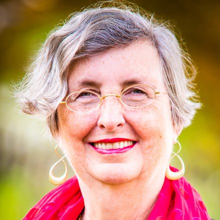 Porträt einer Person mit rotem Schal: Eine Person mit grauen Haaren trägt einen schwarzen Pullover und einen roten, gepunkteten Schal, während sie lächelt.