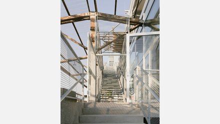 Außentreppe aus verzinktem Stahl mit Gittergeländern; Blick nach oben zwischen Glasfassade und rostigem Dachtragwerk.