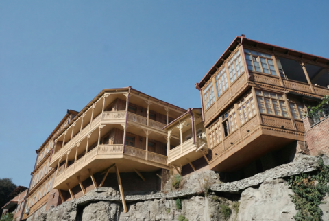Holzgebäude mit aufwändigen, geschnitzten Balkonen und Veranden, das markant über einem steinernen Felsvorsprung vorkragt und von schrägen Holzstützen abgestützt wird; rechteckige Erkerfenster und filigrane Brüstungsornamente prägen die traditionelle Holzfassade, darüber klarer blauer Himmel.