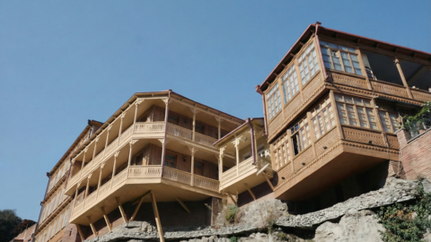 Holzgebäude mit aufwändigen, geschnitzten Balkonen und Veranden, das markant über einem steinernen Felsvorsprung vorkragt und von schrägen Holzstützen abgestützt wird; rechteckige Erkerfenster und filigrane Brüstungsornamente prägen die traditionelle Holzfassade, darüber klarer blauer Himmel.