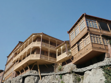 Holzgebäude mit aufwändigen, geschnitzten Balkonen und Veranden, das markant über einem steinernen Felsvorsprung vorkragt und von schrägen Holzstützen abgestützt wird; rechteckige Erkerfenster und filigrane Brüstungsornamente prägen die traditionelle Holzfassade, darüber klarer blauer Himmel.