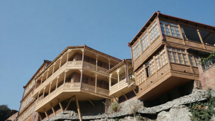 Holzgebäude mit aufwändigen, geschnitzten Balkonen und Veranden, das markant über einem steinernen Felsvorsprung vorkragt und von schrägen Holzstützen abgestützt wird; rechteckige Erkerfenster und filigrane Brüstungsornamente prägen die traditionelle Holzfassade, darüber klarer blauer Himmel.