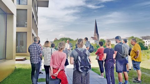 Gruppe von etwa 15 Personen steht auf einem gepflasterten Platz vor einem modernen, cremefarben-grauem Gebäude mit großen Fenstern. Im Hintergrund eine Kleinstadt mit Kirche mit spitzem Turm, grünen Bäumen und bewölktem Himmel. Ein Mann in heller Kleidung scheint die Gruppe anzusprechen, möglicherweise eine Stadtführung oder Informationsveranstaltung.