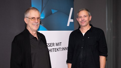 Zwei Männer stehen vor einem Banner, beide lächeln, der linke trägt eine Brille und einen schwarzen Pullover, der rechte ein schwarzes Hemd und helle Hose.