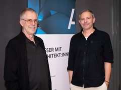 Zwei Männer stehen vor einem Banner, beide lächeln, der linke trägt eine Brille und einen schwarzen Pullover, der rechte ein schwarzes Hemd und helle Hose.