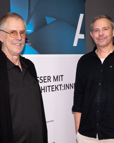Zwei Männer stehen vor einem Banner, beide lächeln, der linke trägt eine Brille und einen schwarzen Pullover, der rechte ein schwarzes Hemd und helle Hose.