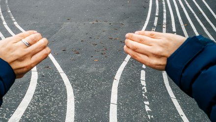 "Vogelperspektive auf einen urbanen Platz mit abstrakt geschwungenen weißen Linien auf dunkelgrauem Asphalt. Im Vordergrund sind die Hände einer Person mit dunklem Ärmel und Ring sichtbar, die auf die Linien zeigen. Die weißen Linien bilden wellenförmige Muster, die sich über den gesamten Platz erstrecken. Im Hintergrund sind ein modernes Gebäude, grüne Vegetation und schwarze Poller sichtbar."