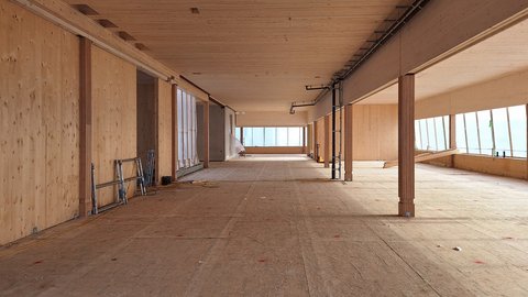 Obergeschoss im Ausbau: Langer offener Raum mit Holzdecke, Holzstützen und Fensterband rechts; provisorischer Boden und etwas Baustellenmaterial.