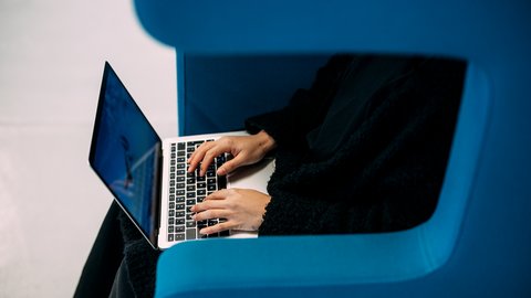 Person sitzt in blauem Sessel und tippt auf Laptop