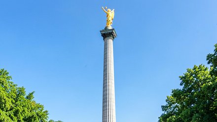 Die Siegessäule mit goldener Victoria oben, hohe zylindrische Säule in einem von Bäumen gesäumten Park, Radfahrer und Spaziergänger am Sockel, klarer blauer Himmel.