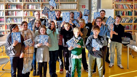 Eine große Gruppe von Kindern und Jugendlichen steht in einer Bibliothek und hält begeistert blaue Bücher mit der Aufschrift „Baukultur“ in die Kamera.