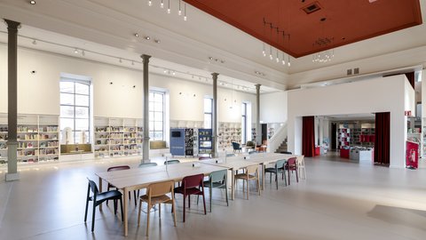 Großer heller Raum mit hohen Fenstern, langen Tischen und bunten Stühlen in einer modernen Bibliothek