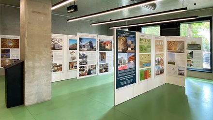 Innenraum einer Ausstellung mit beleuchteten Tafeln zu Holzbauprojekten und Architektur.