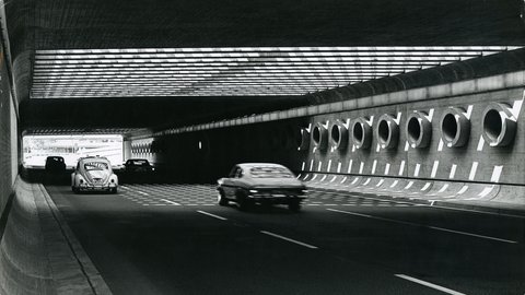 Schwarz-Weiß-Foto aus den 1970ern im Inneren eines brutalistischen Autotunnels. Ein Deckenraster wirft ein Schachbrettmuster aus Licht und Schatten auf die Fahrbahn und die rechte Betonwand, die mit runden Fensteröffnungen versehen ist. Am hellen Tunnelausgang ist Grün zu sehen. Ein heller VW Käfer fährt links, ein anderes Auto überholt rechts mit Bewegungsunschärfe. Die Atmosphäre ist geometrisch und kontrastreich.