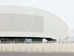 Detailansicht der geschwungenen, weißen Außenfassade des Stadions mit vertikaler Struktur über einer verglasten Erdgeschosszone.