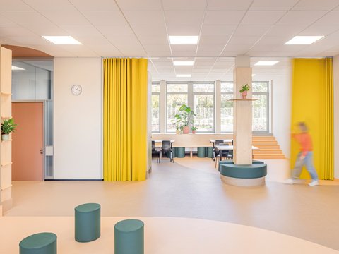"Großzügiger heller Raum mit vielfältigen Nutzungsbereichen. Links orange-rote Türe und ein Kind in Bewegung. Große gelbe Vorhänge auf beiden Seiten zum Abdunkeln. Fensterfront mit Blick ins Grüne. Holztische mit grünen zylindrischen Sitzelementen. Helle Holzböden, breite weiße Pfeiler, indirektes Licht. Pflanzen in rosa und orangefarbenen Töpfen."