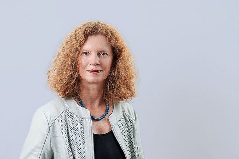 „Porträt einer Frau mit schulterlangem, lockigem rotem Haar. Sie trägt eine helle Jacke über einem schwarzen Oberteil und eine blaue Perlenkette vor hellgrauem Hintergrund.“