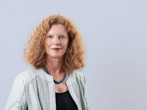 „Porträt einer Frau mit schulterlangem, lockigem rotem Haar. Sie trägt eine helle Jacke über einem schwarzen Oberteil und eine blaue Perlenkette vor hellgrauem Hintergrund.“