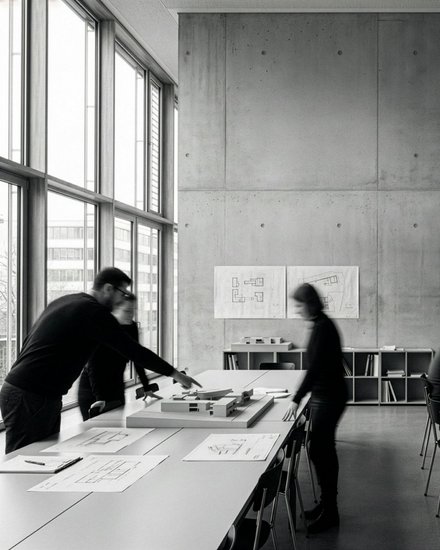 "Modernes Architekturbüro mit Betonwänden und großen Fenstern. Mehrere Personen in dunkler Kleidung arbeiten an weißen Tischen mit Bauplänen und Laptops. An der Wand hängen Grundrisse und eine digitale Anzeige zeigt ein 3D-Architekturmodell eines Wohngebäudes. Regale mit Fachbüchern, Schreibmaterialien und Modelle im Hintergrund.