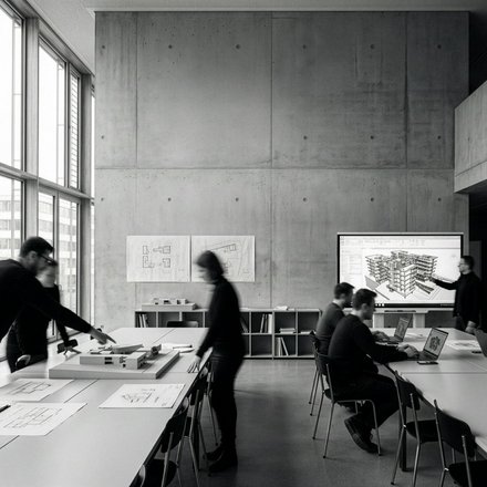 "Modernes Architekturbüro mit Betonwänden und großen Fenstern. Mehrere Personen in dunkler Kleidung arbeiten an weißen Tischen mit Bauplänen und Laptops. An der Wand hängen Grundrisse und eine digitale Anzeige zeigt ein 3D-Architekturmodell eines Wohngebäudes. Regale mit Fachbüchern, Schreibmaterialien und Modelle im Hintergrund.