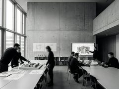 "Modernes Architekturbüro mit Betonwänden und großen Fenstern. Mehrere Personen in dunkler Kleidung arbeiten an weißen Tischen mit Bauplänen und Laptops. An der Wand hängen Grundrisse und eine digitale Anzeige zeigt ein 3D-Architekturmodell eines Wohngebäudes. Regale mit Fachbüchern, Schreibmaterialien und Modelle im Hintergrund.